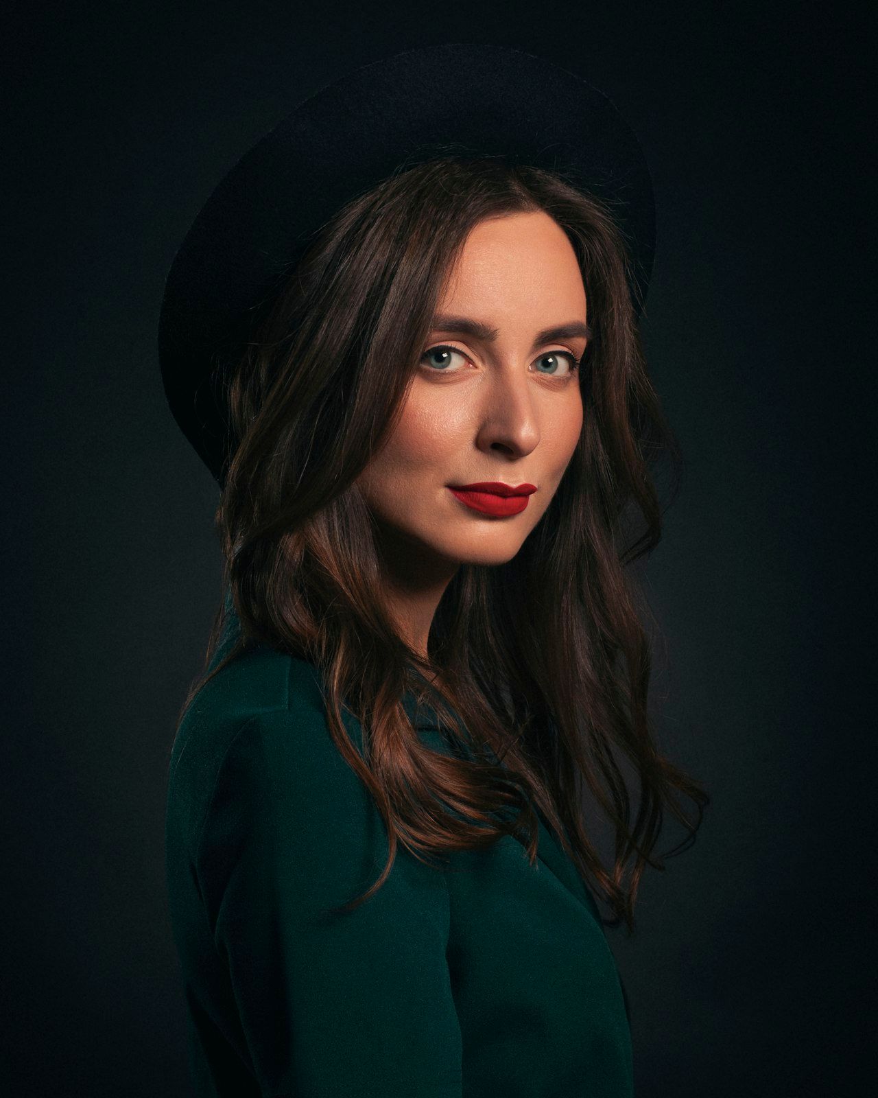 A portrait in a wide black hat and green coat against deep shadow, red lip.