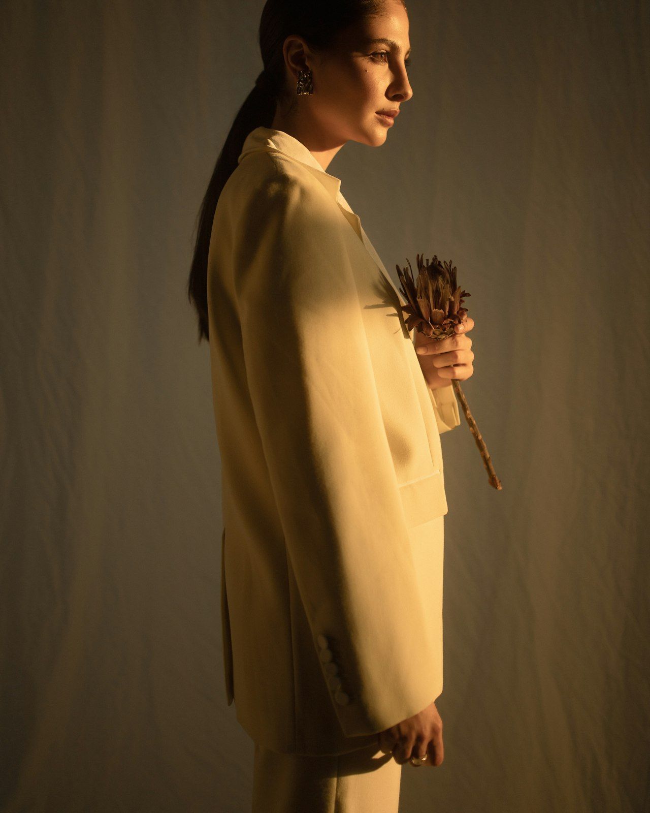 A model in profile wearing a cream suit, holding a dried protea in low warm light.