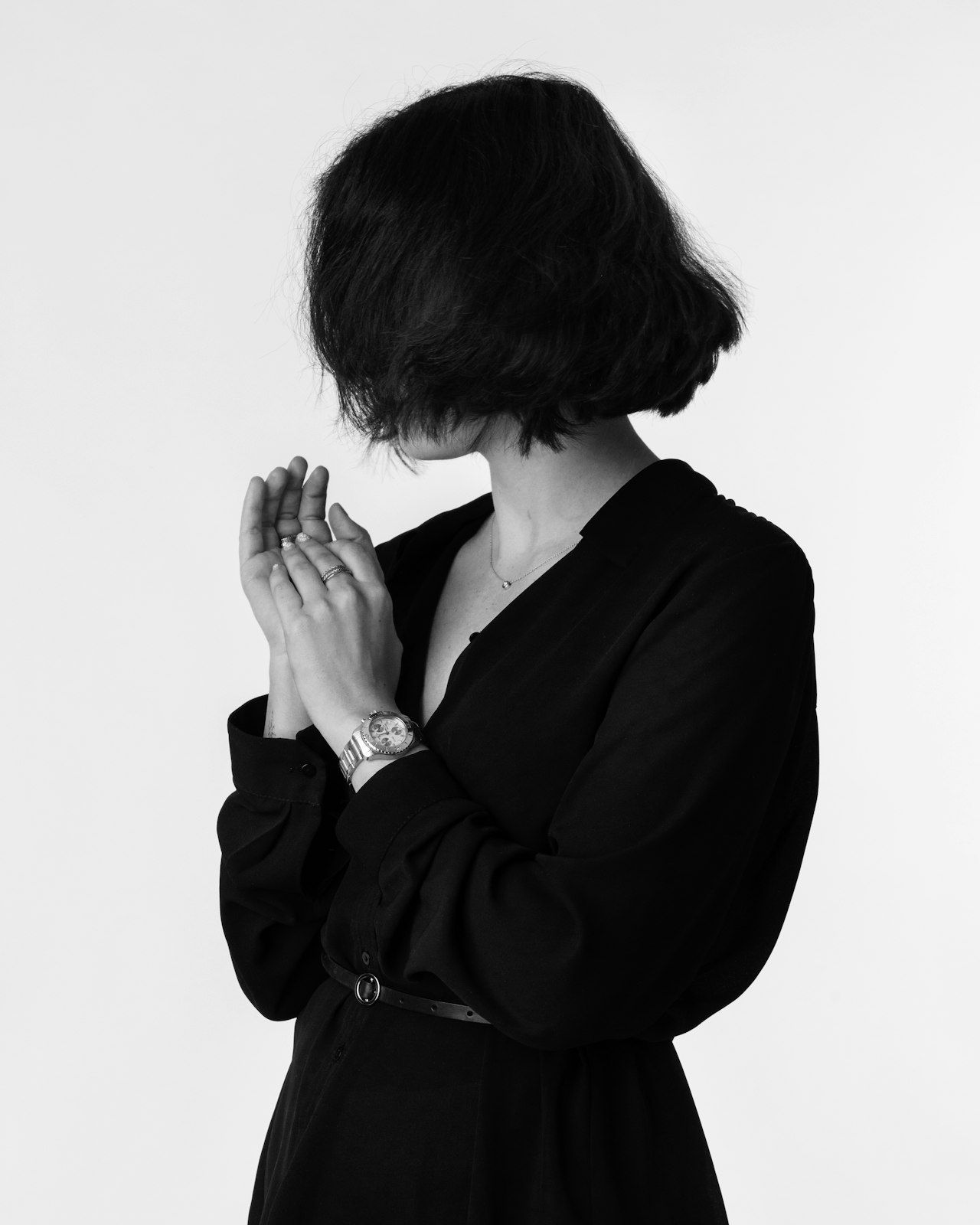 A black-and-white figure in a dark dress hiding her face behind clasped hands.